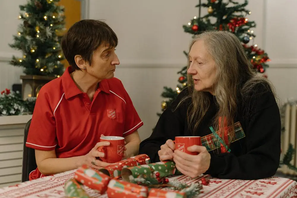 Salvation Army staff member and an older woman chatting over hot drinks at a community Christmas lunch. Models used.
