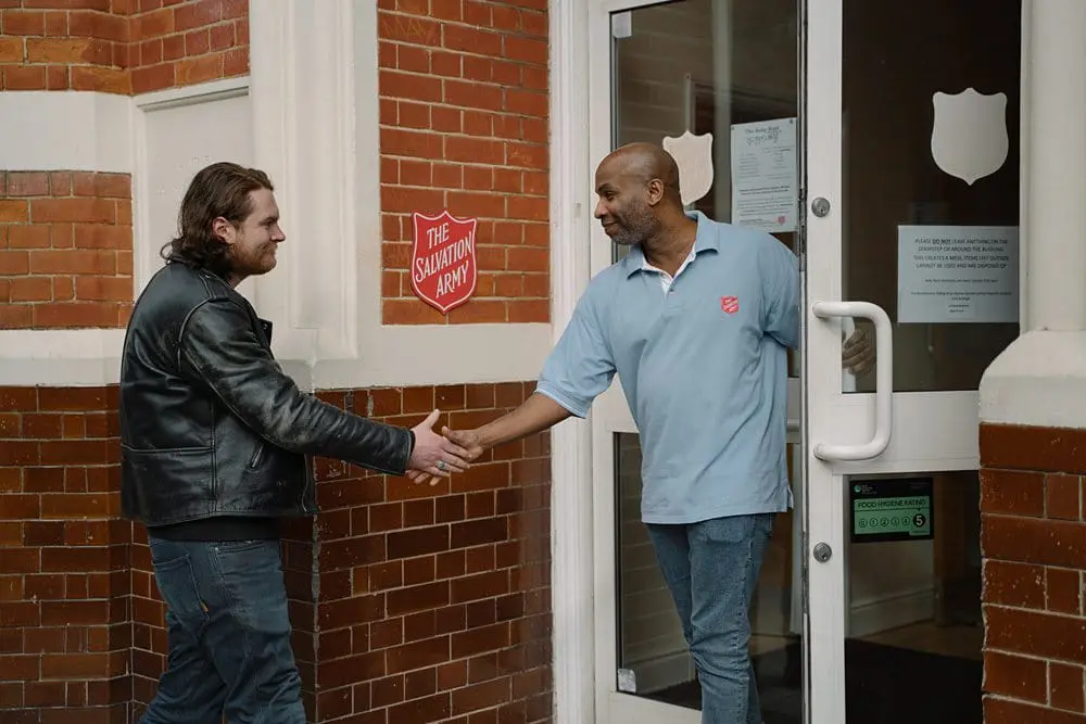 Salvation Army staff member warmly shaking hands with a visitor at the entrance of a community centre. Models were used.