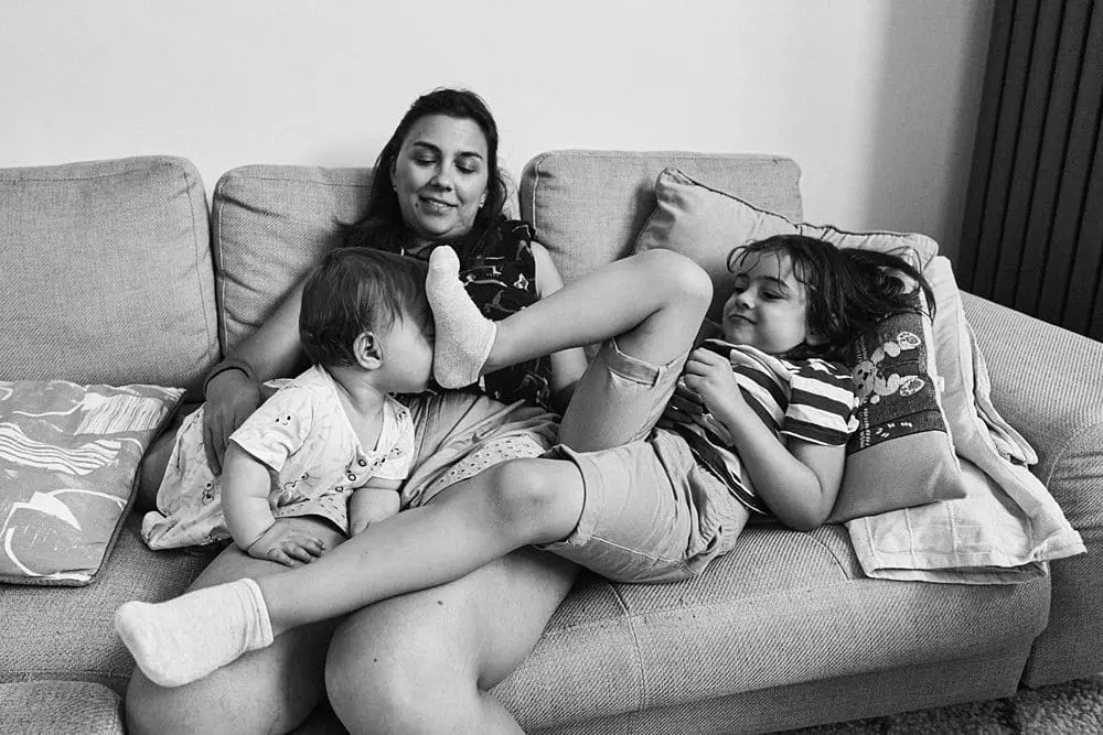 Mum on the sofa with her baby and her older child stretched across her lap — relaxed at-home family photography Blackheath
