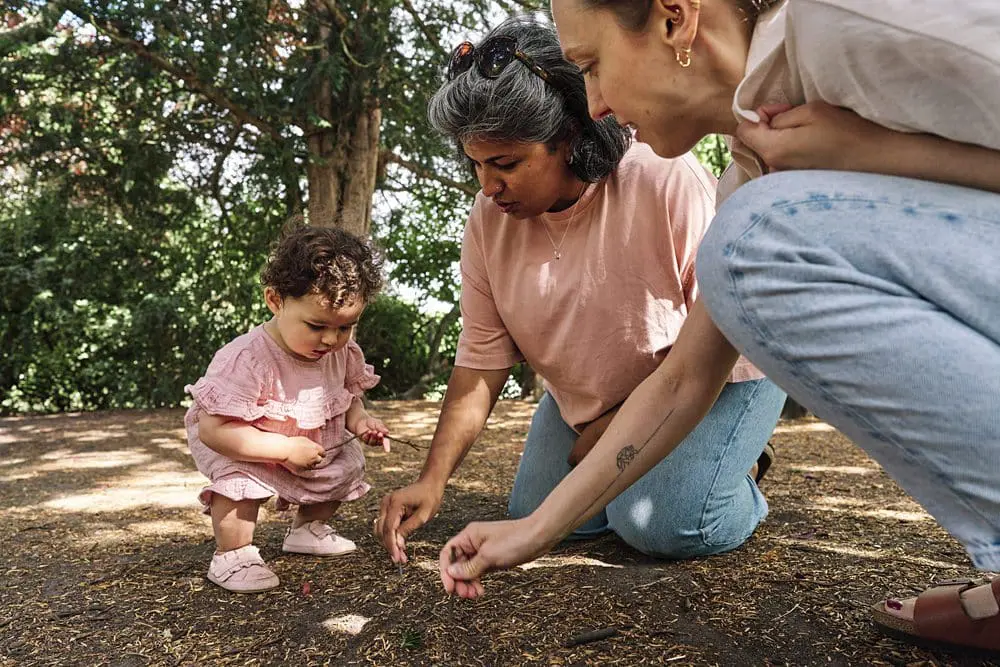 Young child exploring and drawing with sticks in sand with her mums — candid family photography South-East London