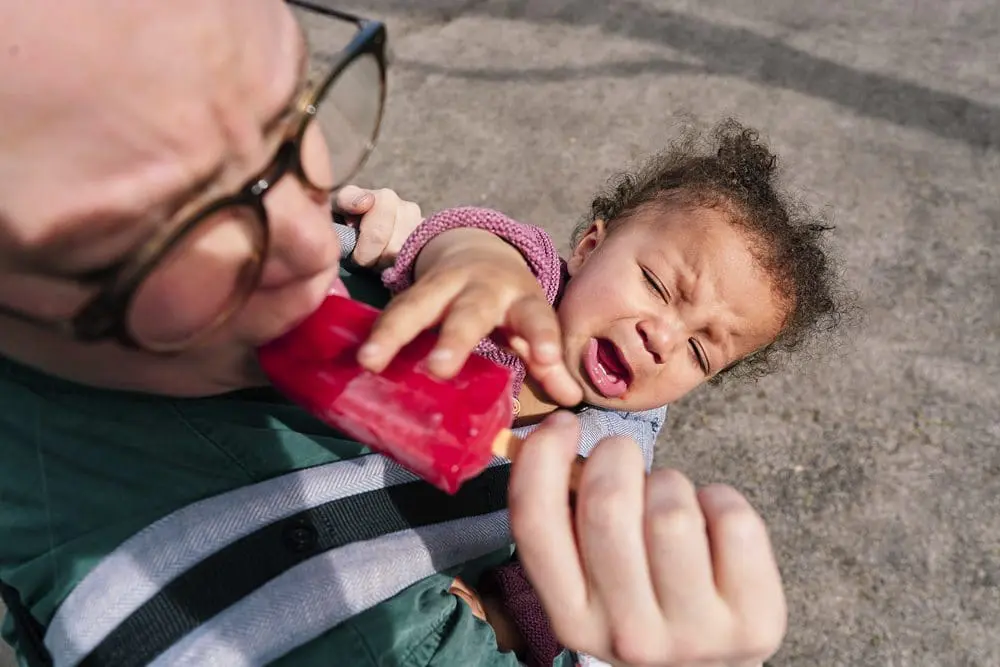 Toddler in a sling reaching out to grab an ice cream — playful documentary family photography Greenwich SE10