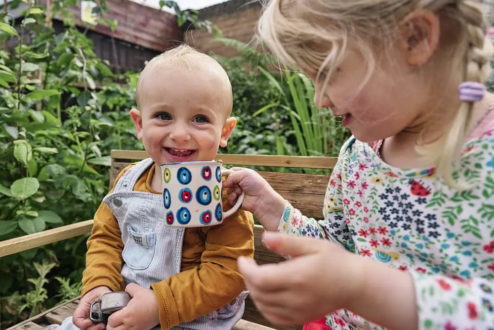 Child smiling naturally as sister helps him drink from a cup during a documentary family photo session in London