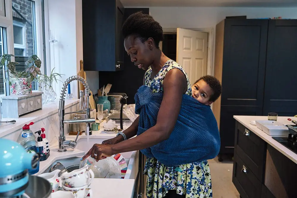 Mum washing up in the kitchen with her baby in a sling on her back during a documentary family photo session at home