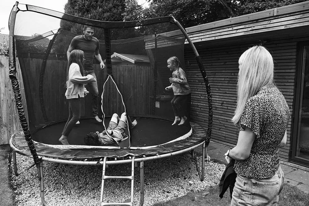 Father and children jumping on a trampoline as mother watches in a garden in Surrey — relaxed family photography South-East London