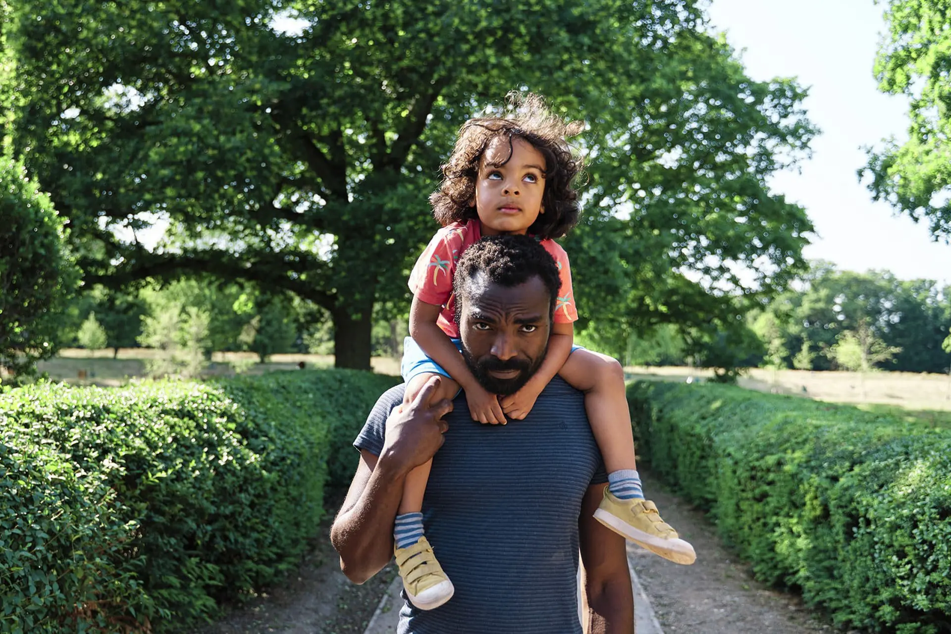 Dad carrying his daughter on his shoulders along a hedge-lined park path, an easy outdoor idea for a one-hour mini session