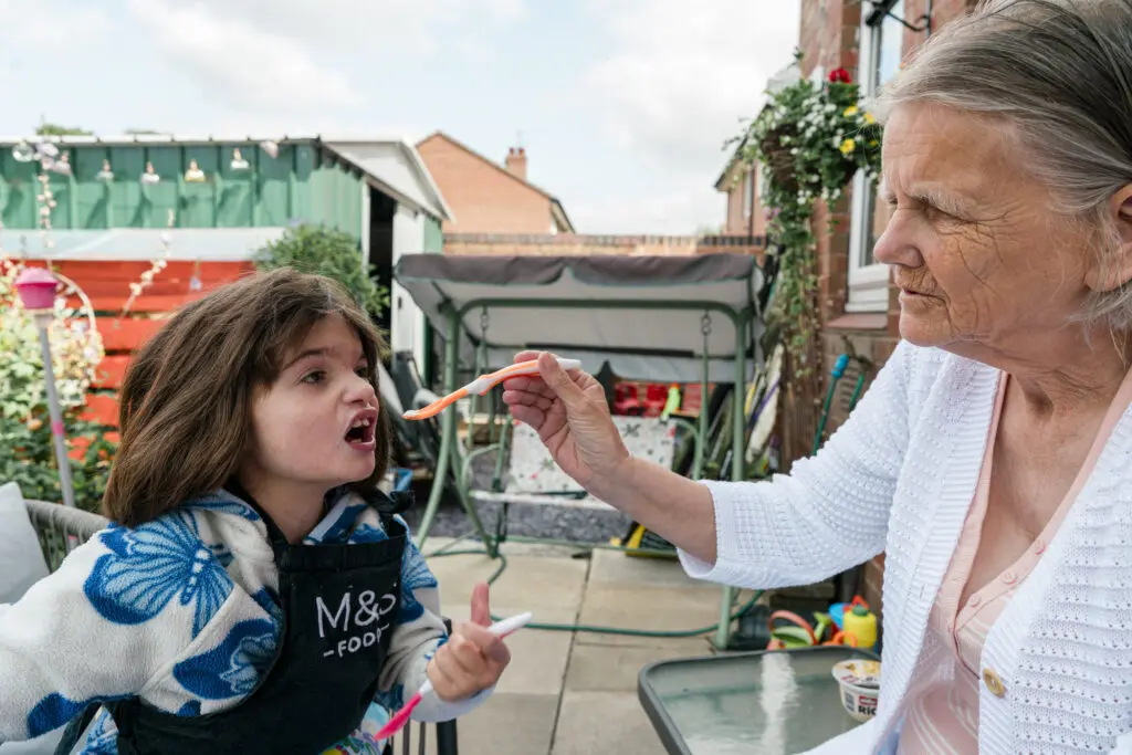 Grandmother and kinship carer gently feeding her granddaughter in the back garden, real documentary charity photography rather than stock images