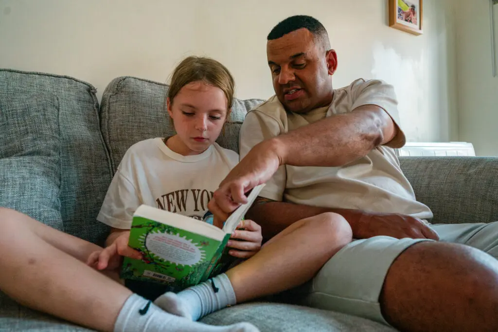 Girl reading a book to her uncle and kinship carer on the sofa at home — documentary photography for Kinship Care Week