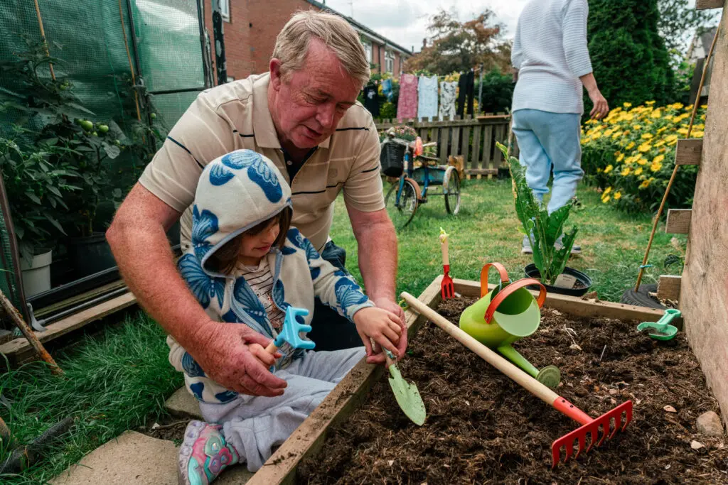 Grandfather and kinship carer planting seeds in a raised garden bed with his young granddaughter — documentary photography for charities