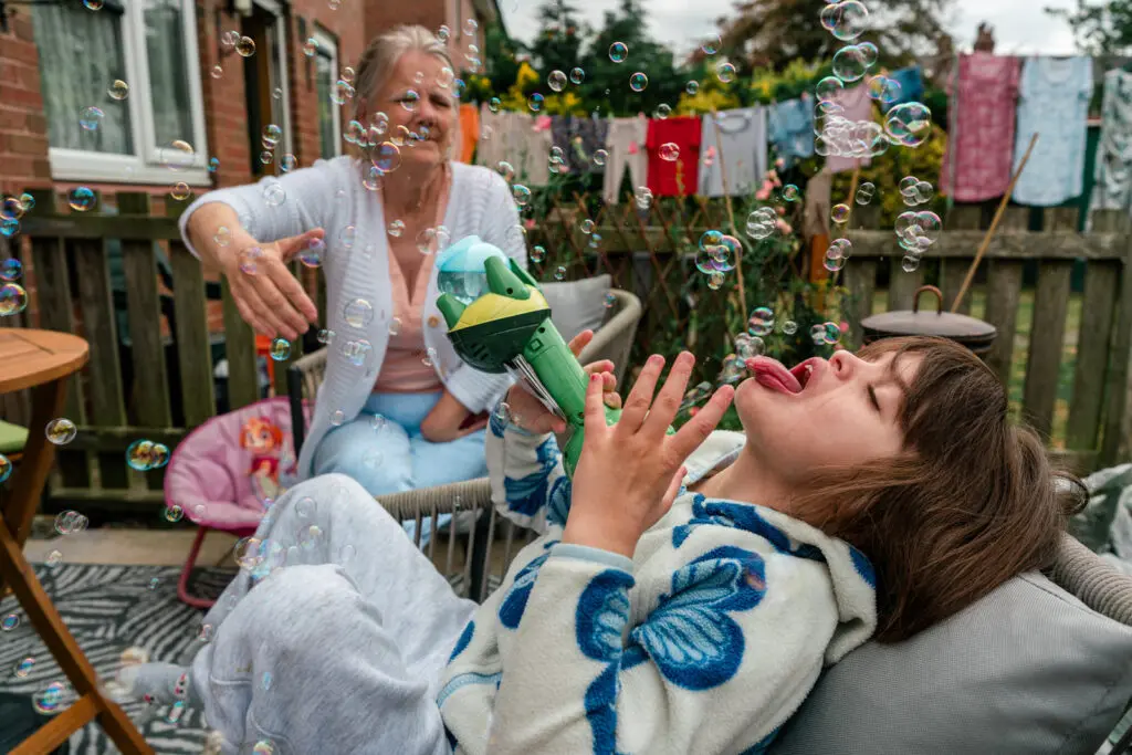 Kinship carer playing with bubbles in the back garden while the granddaughter leans back to catch them on her tongue — documentary charity photography for Kinship