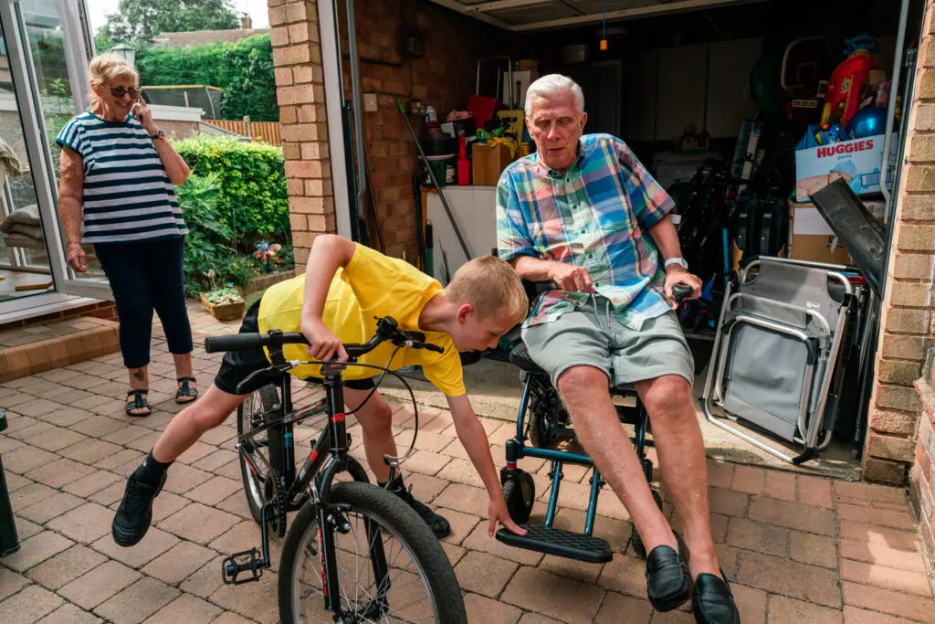 Boy on his bike leaning over to help push his grandfather's wheelchair out of the garage as his grandmother looks on — kinship family documentary photography