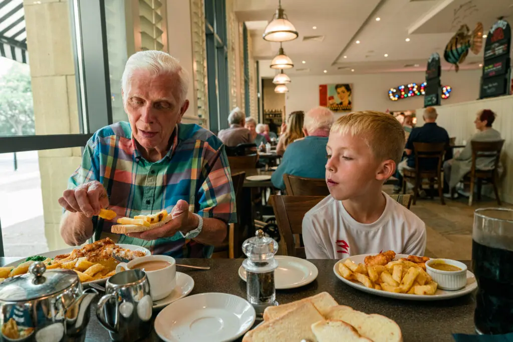 Grandfather showing his grandson how to make a chip butty at a café table — storytelling charity photography