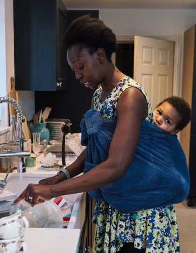 Family Photographer and filmmaker in London photographing a mother with baby on her back doing the dishes.