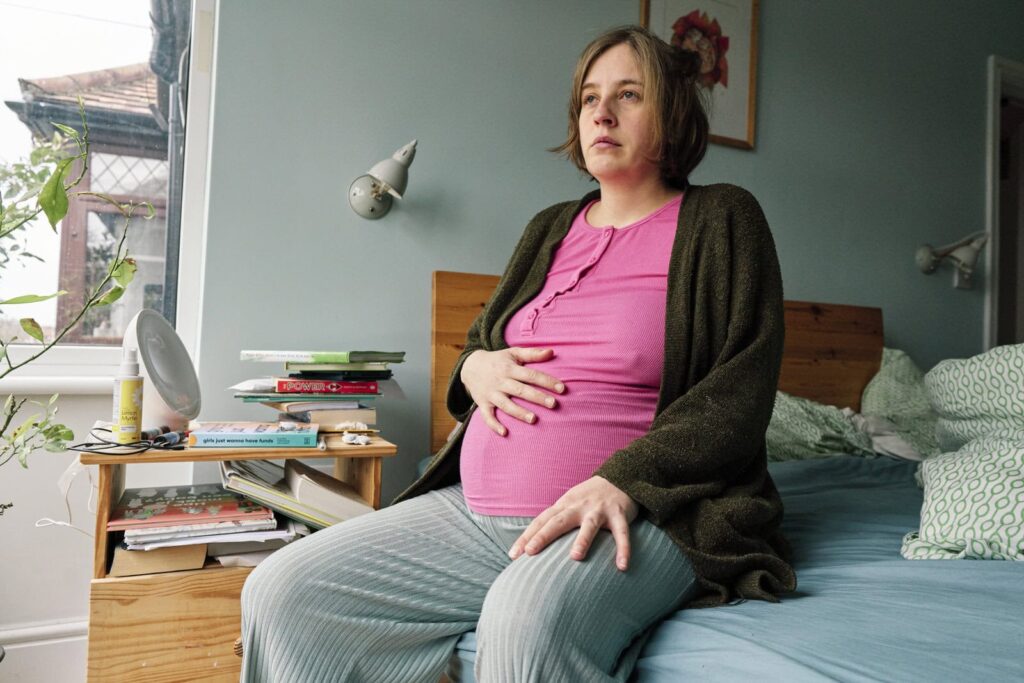 Pregnant woman in pyjamas sitting on the bed – documentary-style maternity photography.