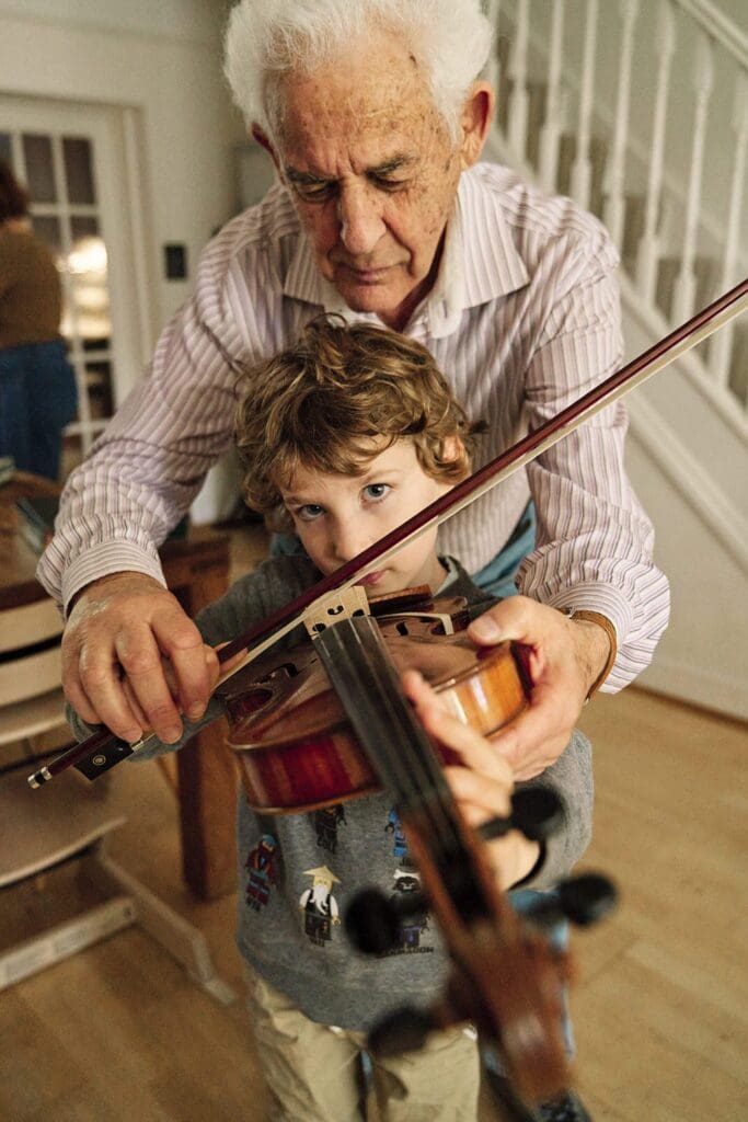 Boy looking into the camera of the photographer as his grandfather helps him play violin — tender multi-generational documentary family photography London